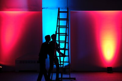 Innenraum in rotem und mittig in blauem Licht erleuchtet, in der Mitte zwei Personen als Umrisse eine Leiter verstellend
