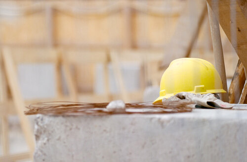 ein gelber Arbeitsschutzhelm liegt auf einem Betonblock, im Hintergrund Holzbauteile auf einer Baustelle