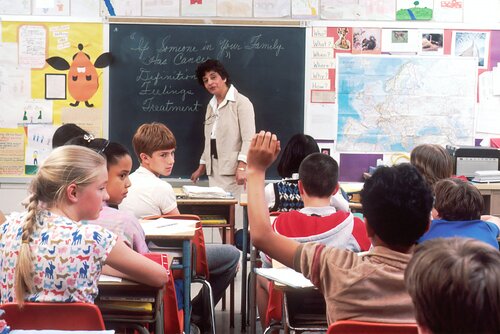 Grundschüler im Klassenraum sitzend blicken auf einen Schüler, Lehrerin steht an der Tafel