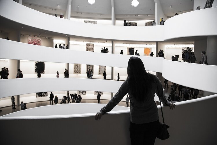 Wendeltreppe im Museum, kühle Farben im Weiß-grau-Modus, im Vordergrund steht eine Frau am Treppengeländer und schaut in den Ausstellungsraum