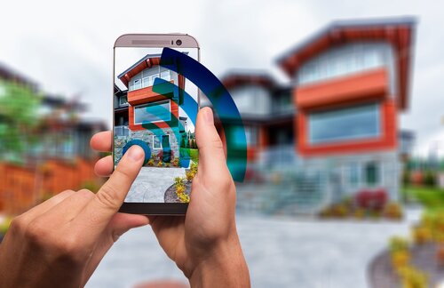 Smarthome: eine Hand bedient im Vordergrund eine Smartphone-App, im Hintergrund ein Haus