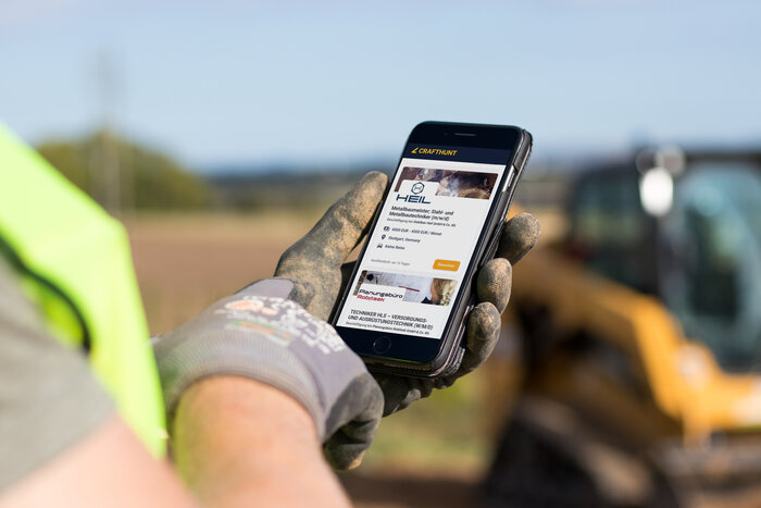 ein Mensch mit Arbeitsschutzweste auf einer Baustelle hält ein Smartphone in der Hand, auf dem "BauGPT" aufgerufen ist