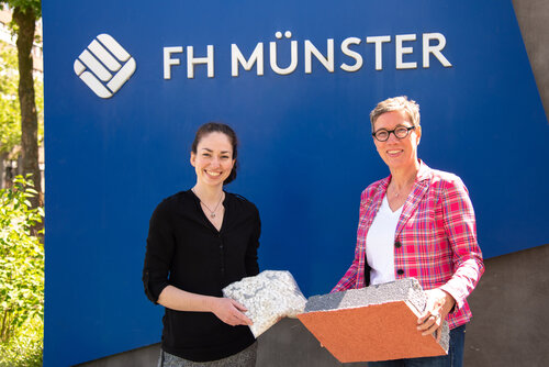 Jana Winkelkötter (v. l.) und Prof. Dr. Sabine Flamme von der FH Münster vor einer blauen Fläche mit Logo der FH Münster, aufgenommen vor der FH