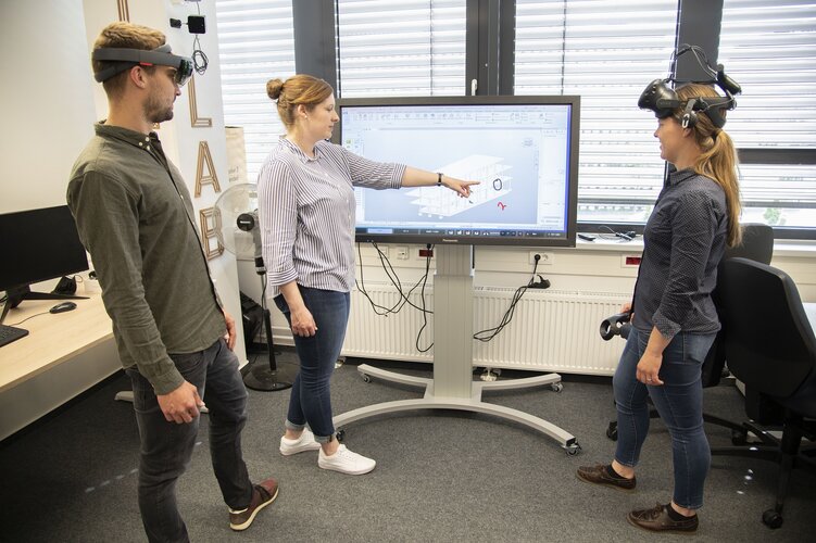 Wissenschaftler:innen stehen im Büro vor einem Monitor und betrachten ein digitales Gebäudemodell, zwei Forscher tragen eine VR-Brille auf dem Kopf