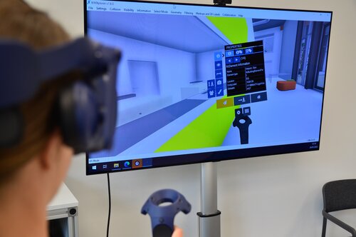Digitales Gebäudemodell auf Monitor, davor Hinterkopf einer Person mit Virtual-Reality-Brillen
