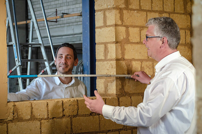 Der Technische Berater, Stefan Dubelzieg (r.), und Vertriebsmitarbeiter Sascha Möller stehen an einer Lehmsteinwand und legen bei der Prüfung des Lehm-Mauerwerks selbst Hand an.